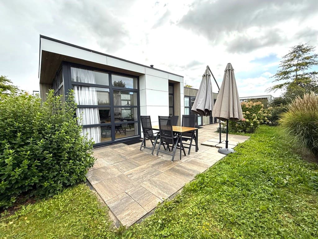 Moderne vakantiewoning met betegeld terras, tuinmeubilair en parasols in groene omgeving.