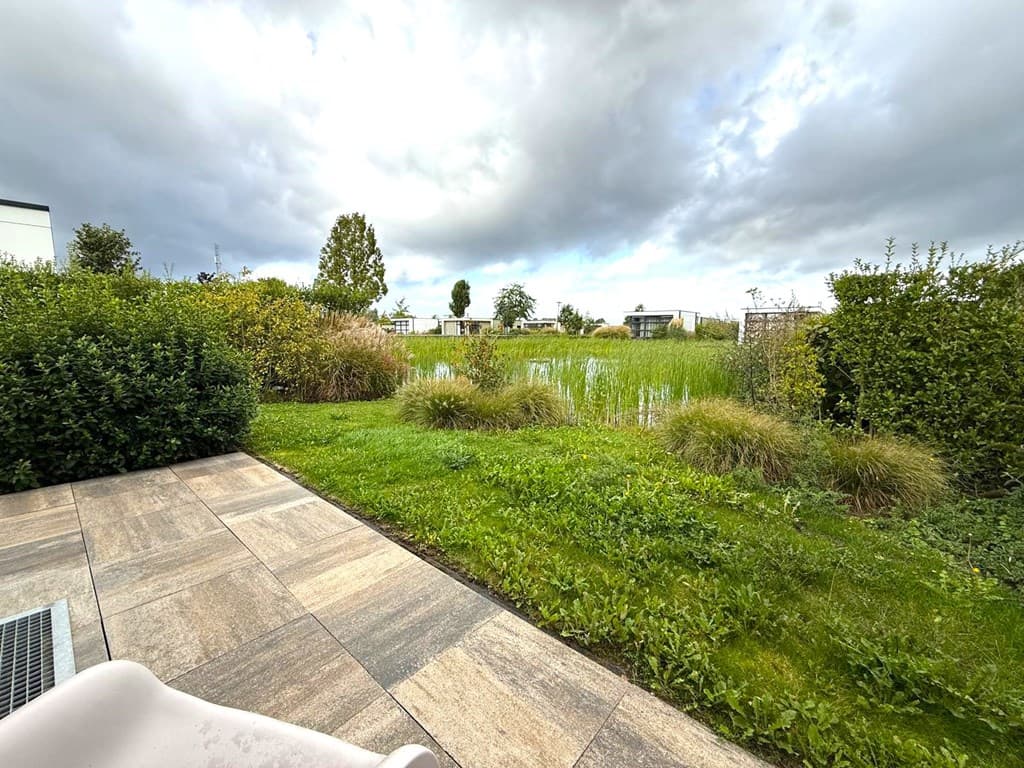 Uitzicht vanaf een terras met gras en struiken, kijkt uit over een groene tuin met riet en vakantiehuizen op de achtergrond