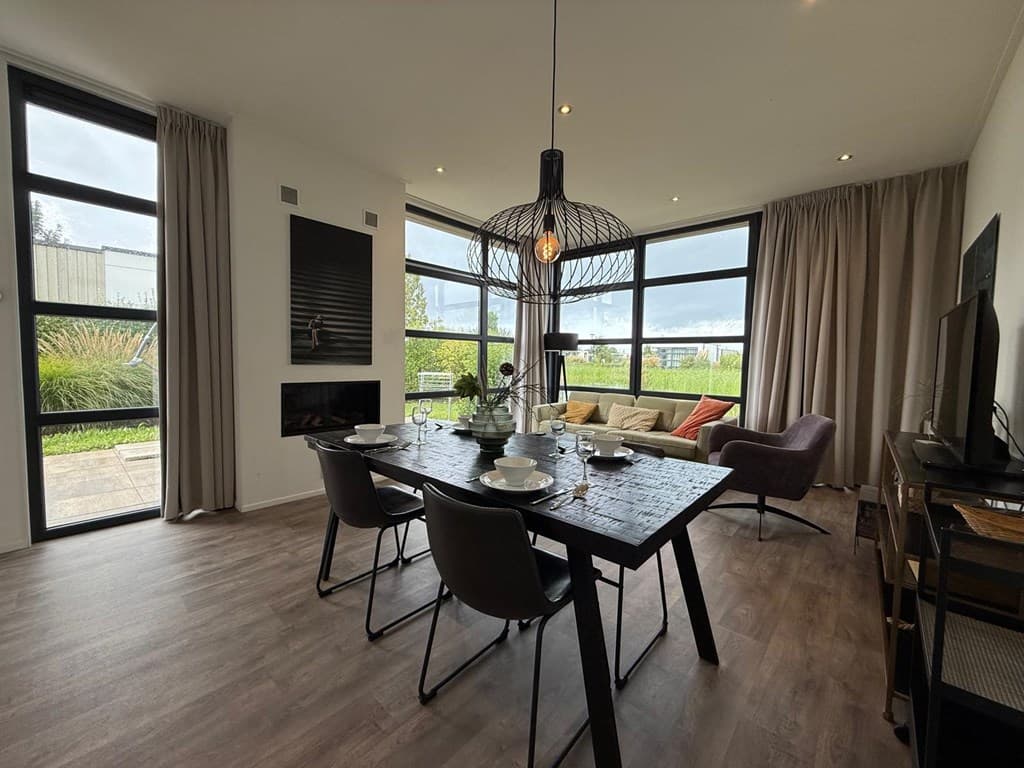 Moderne woonkamer met grote ramen, een houten eettafel en een zithoek met gezellige sofa en fauteuils