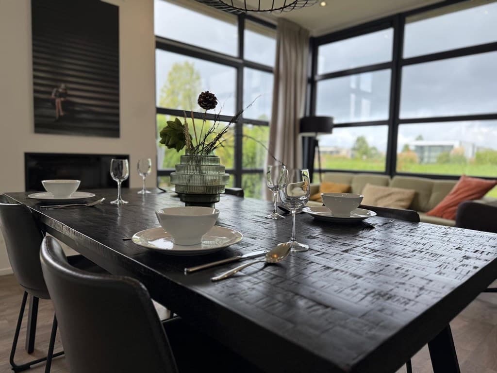 Eetkamer met een grote zwarte tafel, witte borden en glazen, en volop natuurlijk licht door grote ramen.