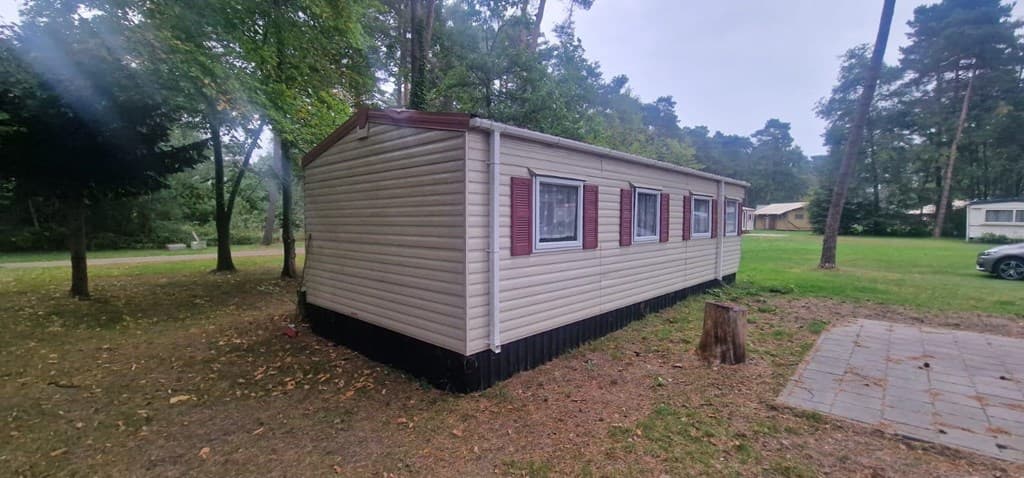 Zijaanzicht van een vakantiewoning met houten omheining en grasveld in de omgeving van bomen en andere woningen.