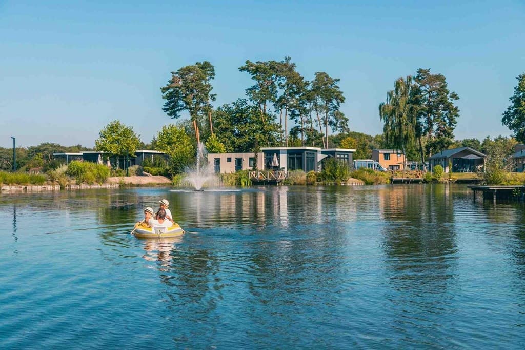 Twee mensen op een gele pedalboot op rustig water omringd door groene bomen en vakantiewoningen in de verte.