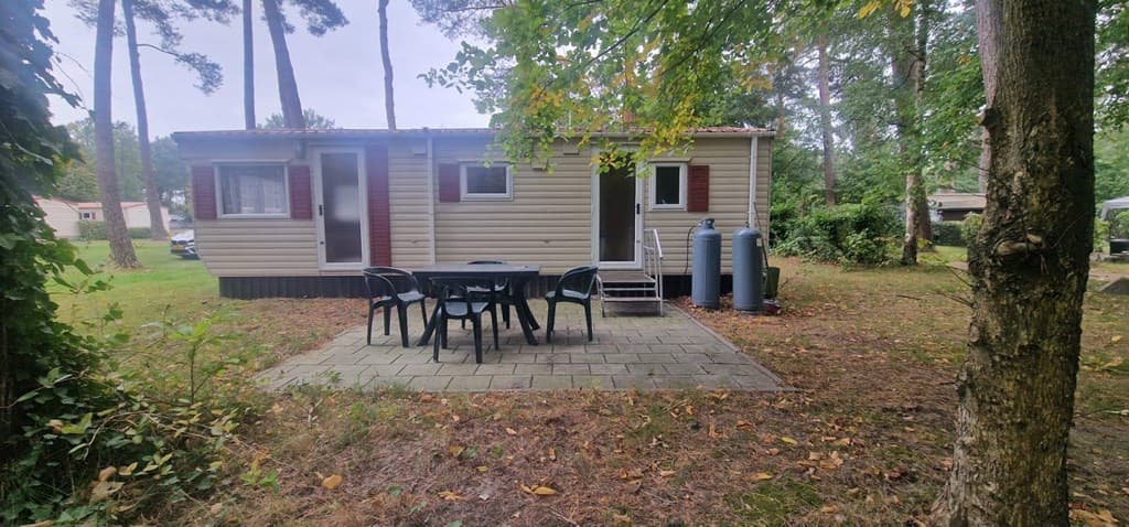 Houten vakantiewoning met terras en tuinmeubilair in een bosrijke omgeving met bomen rondom.