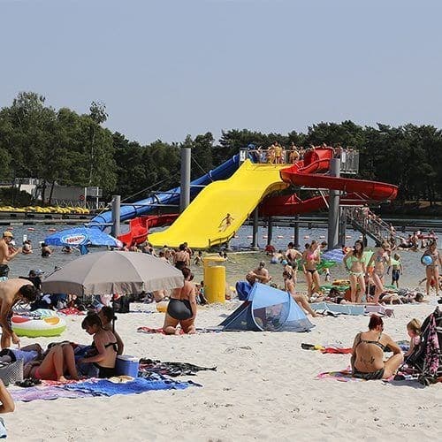 Zandstrand met veel mensen, parasols en kleurrijke glijbanen aan het water van EuroParcs Zilverstrand.