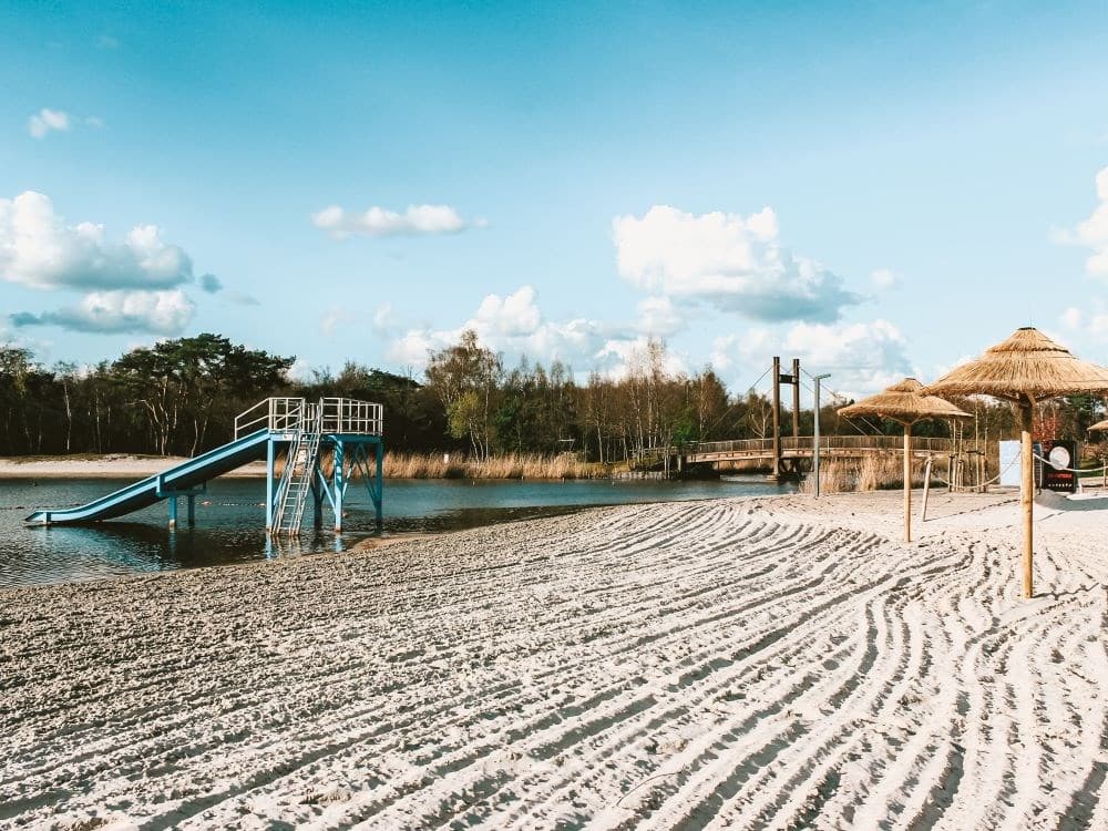 Zicht op een glijbaan aan het water, met zandstrand en parasols onder een blauwe lucht met wolken