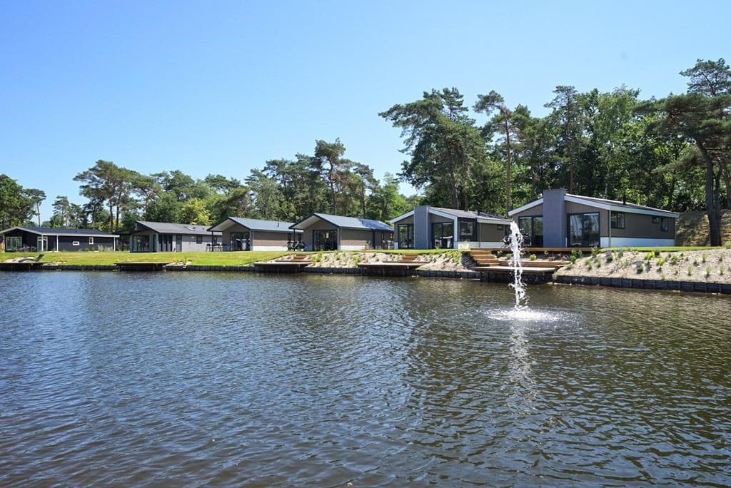 Zicht op moderne vakantiewoningen aan een meer met een fontein en groene bosachtige omgeving