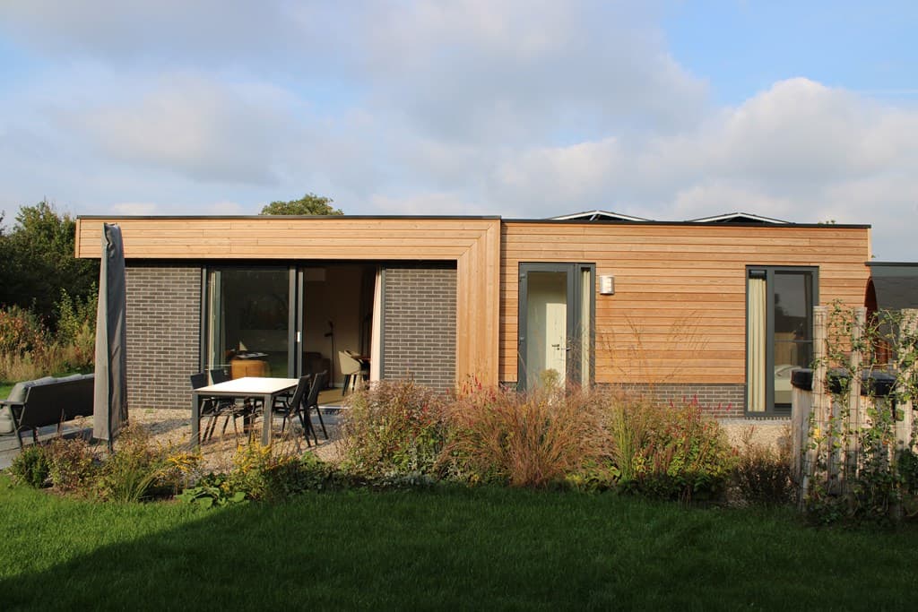 Moderne vakantiewoning met houten gevel en grote ramen, omringd door een tuin met gras en planten.