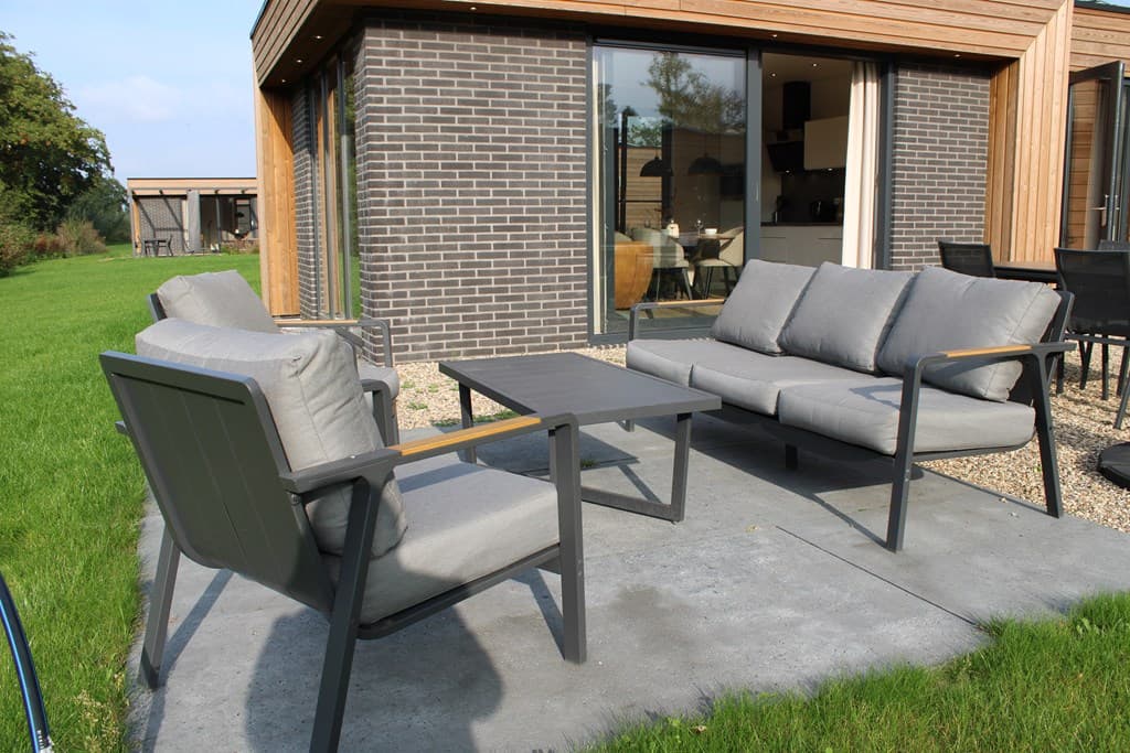 Tuin terras met een zithoek van grijze tuinstoelen en een lage tafel, gelegen op een betegelde ondergrond.