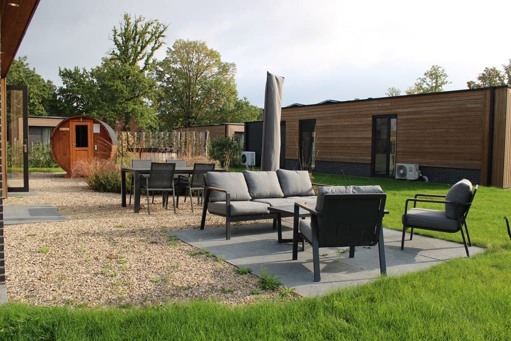 Tuin met loungemeubels, een eettafel en een houten sauna in een vakantiepark met groene grasvelden en bomen.