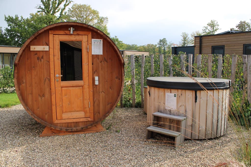 Houten sauna in de vorm van een vaten met een hot tub ernaast, omringd door planten en een houten hek.