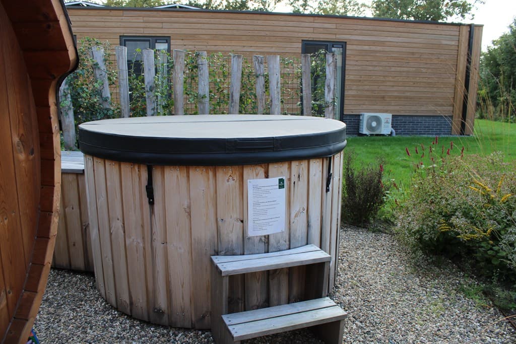 Houten wellnessbad met een trapje, omringd door een groene tuin en een moderne woning op de achtergrond