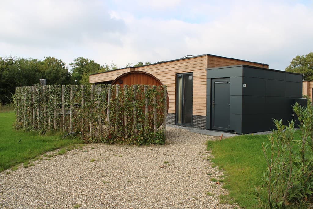 Moderne vakantiewoning met houten gevel en grijze aanbouw, omgeven door een omheining van hout en groen, gelegen in een parka