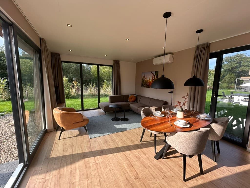 Moderne woonkamer met grote ramen, bank, eetgedeelte en uitzicht op groene omgeving in vakantiepark Hofparken Wiltershaar.