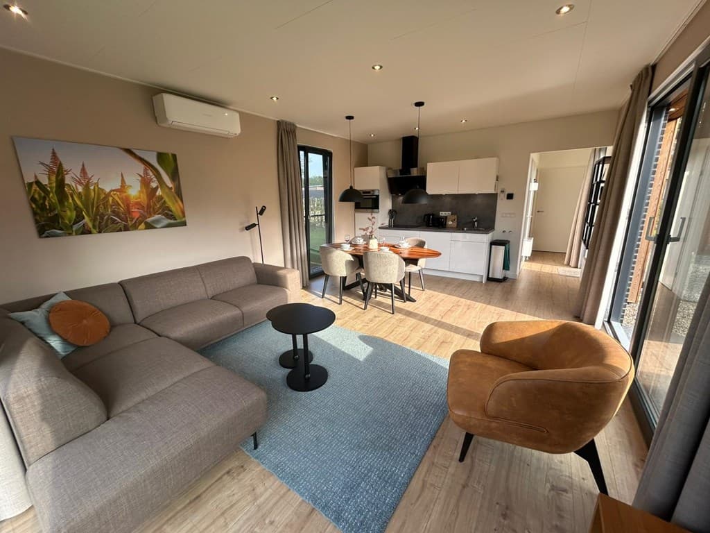 Moderne woonkamer met bank, eettafel en open keuken, grote ramen met uitzicht op buitenruimte en planten aan de muur.
