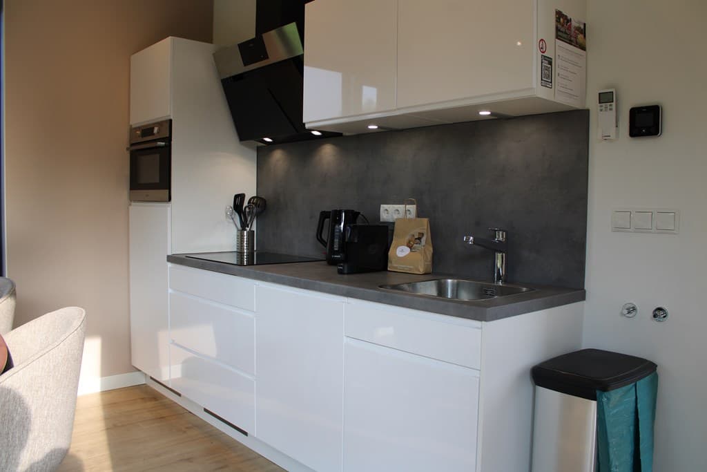 Moderne keuken met witte kasten, spoelbak, kookplaat en verschillende apparatuur aan een grijs aanrecht.