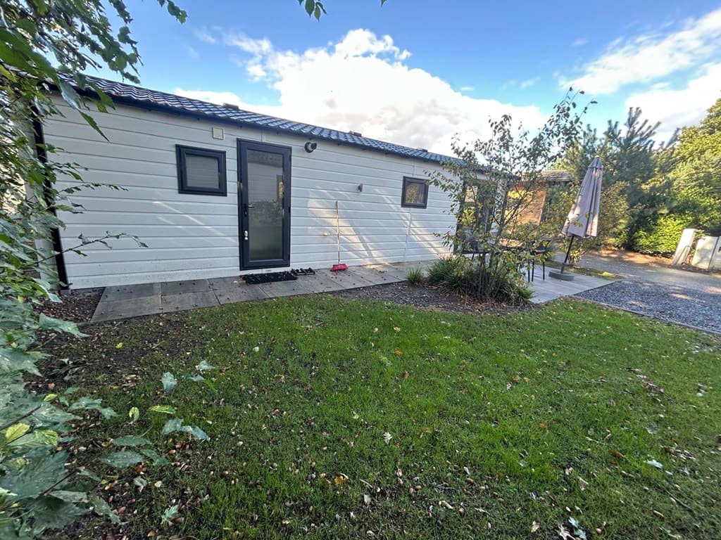 Wit vakantiehuisje met een grasveld en terras, omringd door bomen en struiken in een rustige omgeving.