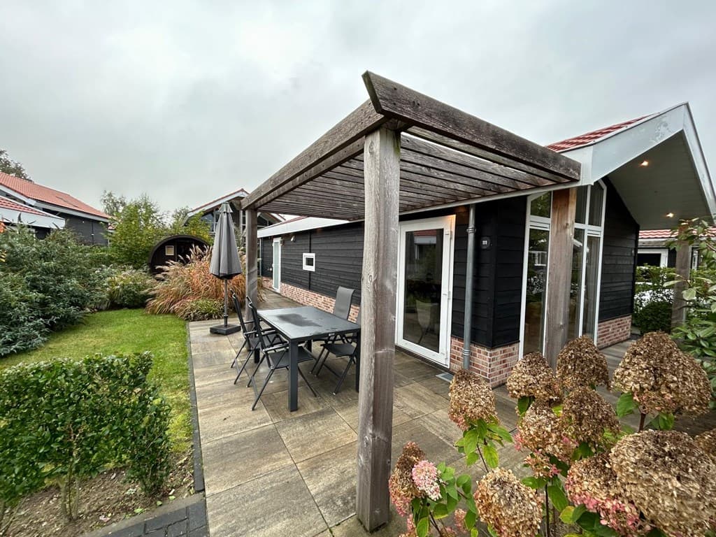 Zwarte vakantiewoning met veranda, eettafel en stoelen, omgeven door groen en bloemen in de tuin