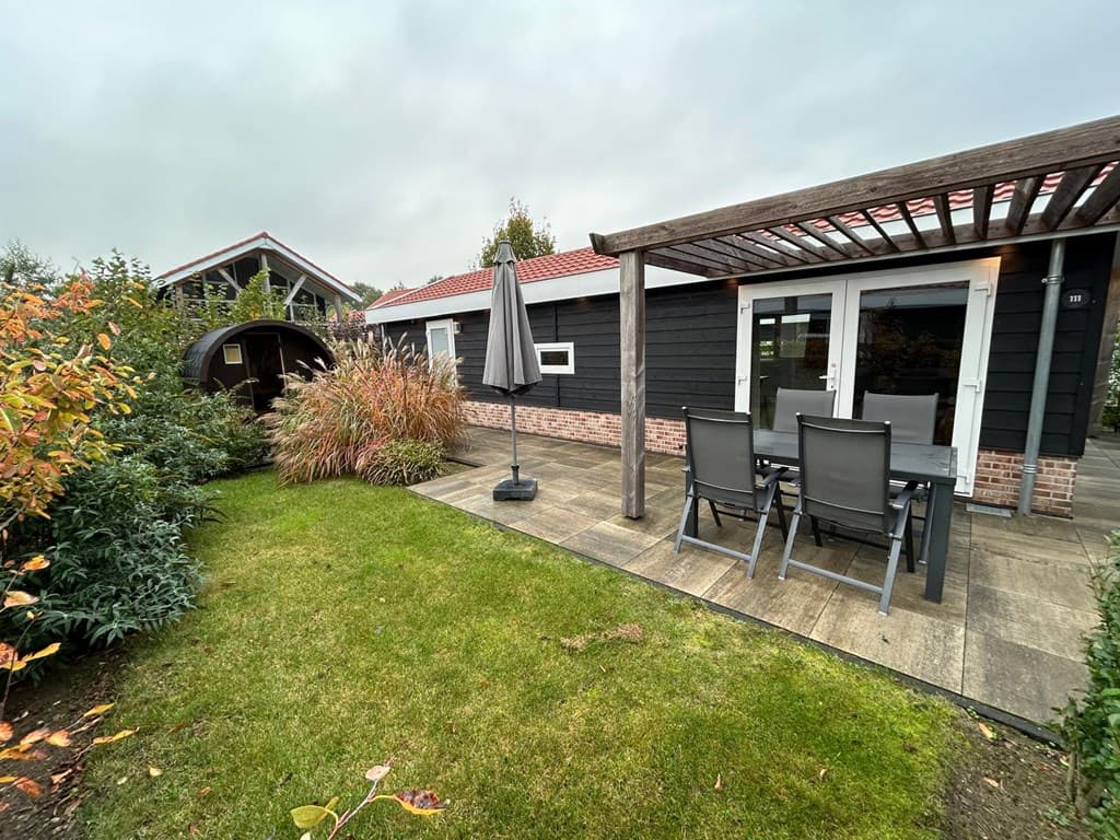 Tuin met terras, zes tuinmeubels en een parasol, omringd door groen en een houten huisje aan de achterkant