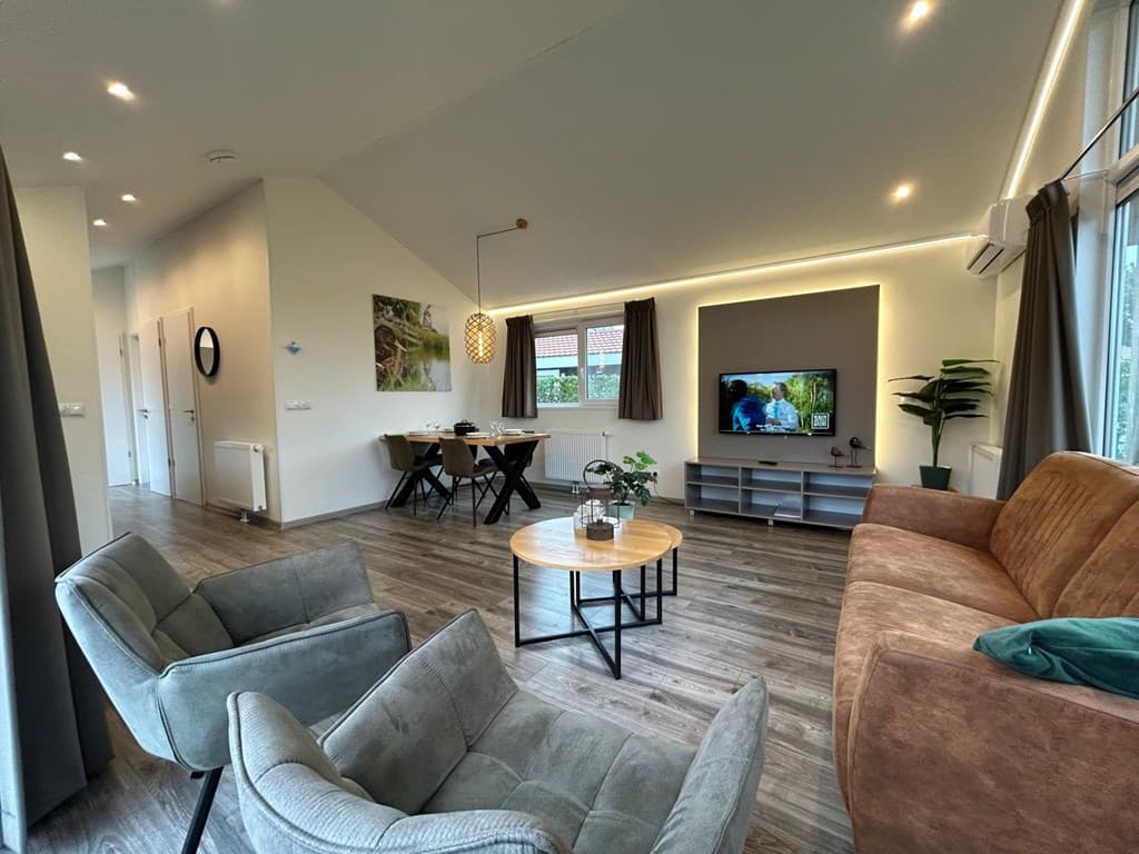 Moderne woonkamer met zithoek, eettafel en televisie, grote ramen en sfeervolle verlichting.