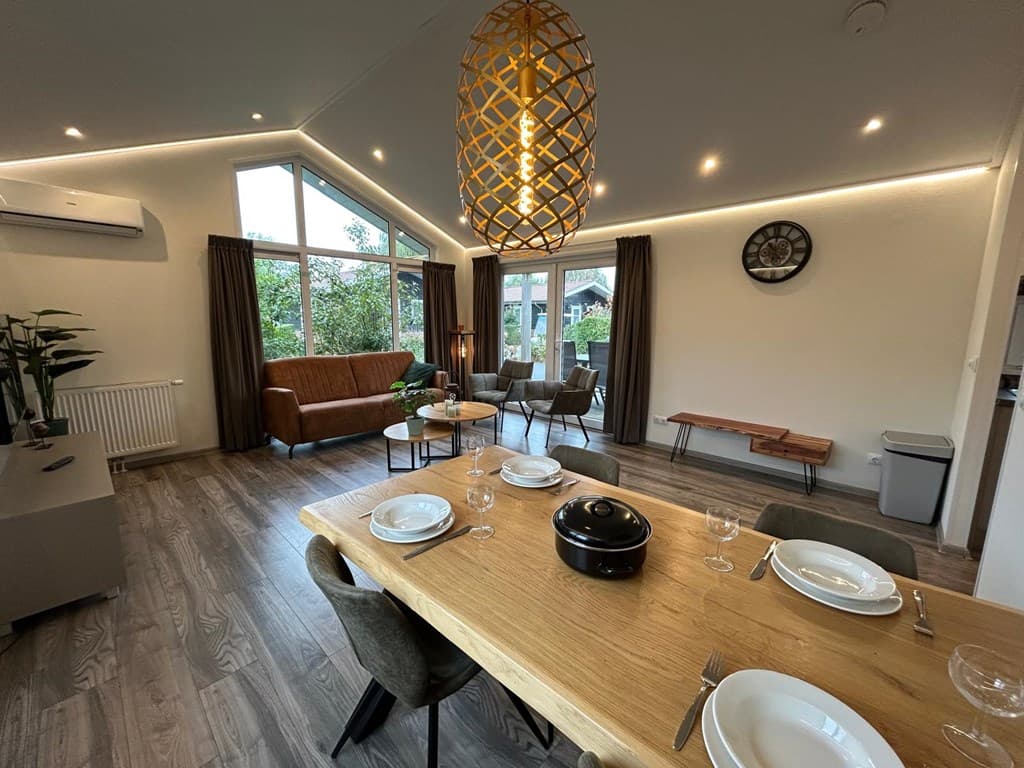 Eetkamer met houten tafel, glazen servies en comfortabele zitbank met fauteuils en grote ramen voor natuurlijk licht.