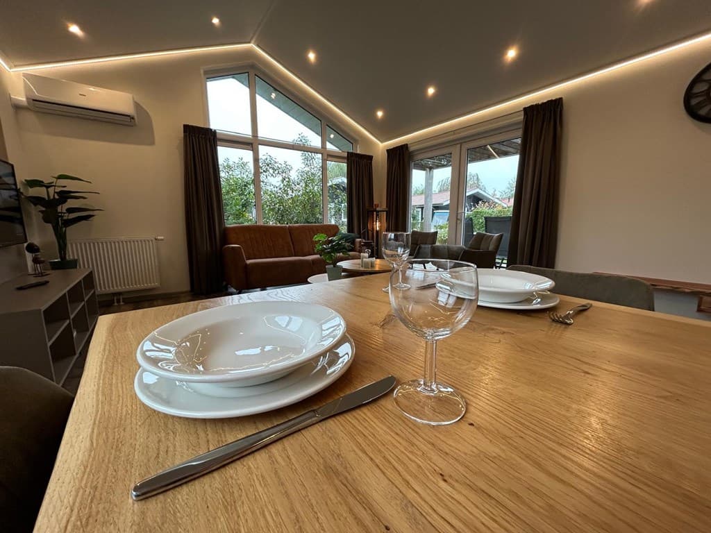 Interieur met eettafel, servies en uitzicht op zithoek bij ramen met natuurlijk licht en groene omgeving.
