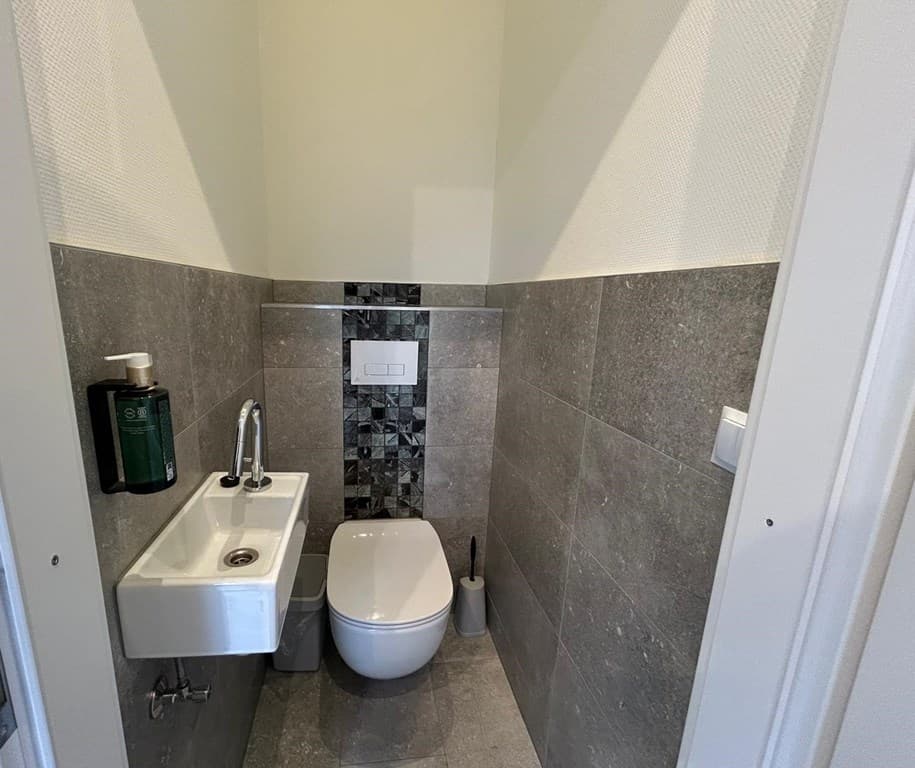 Toilet met wanden van grijze tegels en een kleine wastafel met kraan, netjes en modern ingericht.