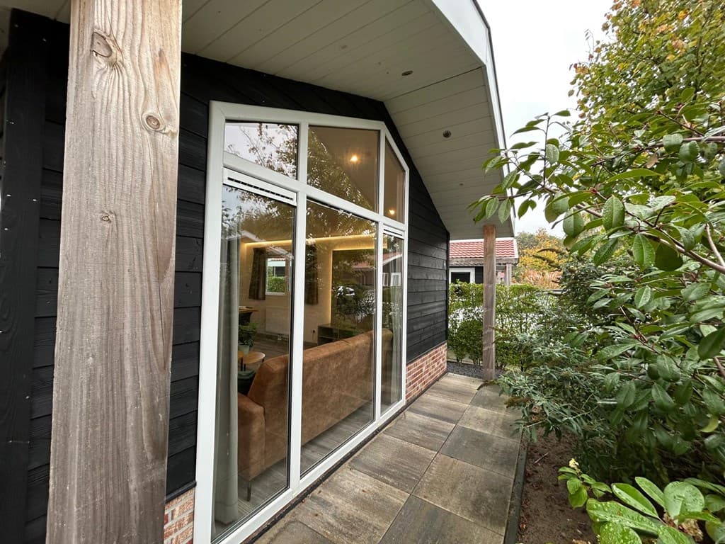 Entree van een vakantiehuis met grote ramen en een betegeld pad, omringd door groene planten en bomen