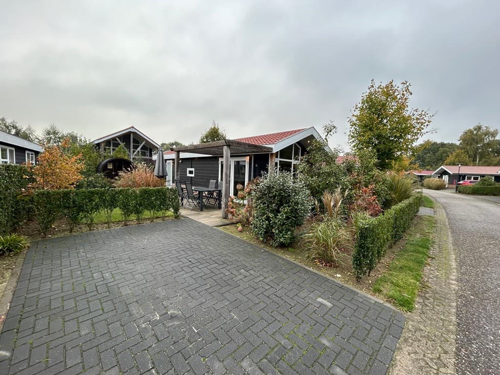 Zicht op een vakantiewoning met een betegelde oprit, groene hagen en kleurrijke beplanting, gelegen aan een rustige straat.