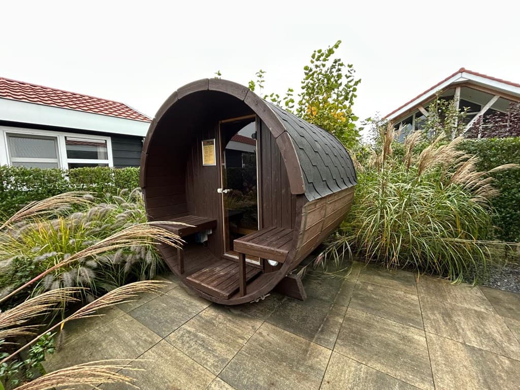 Houten sauna in de tuin met een ronde vorm en bankjes aan de zijkanten, omringd door groen en plantengroei