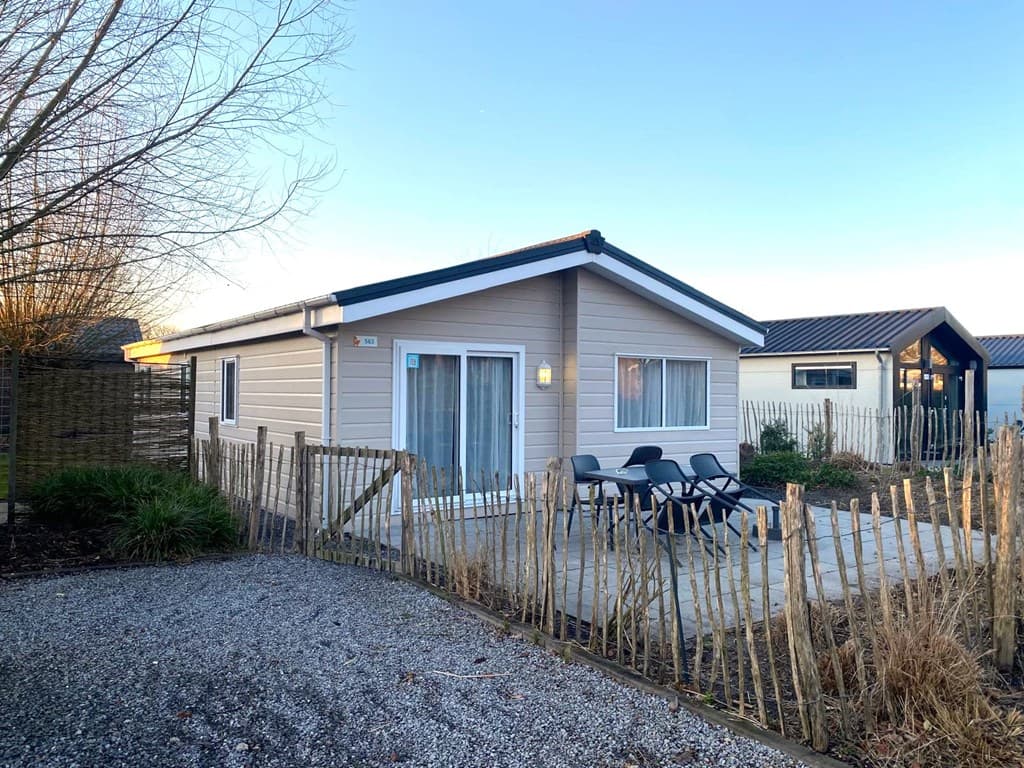 Moderne vakantiewoning met glazen schuifdeur, terras en zitmeubelen, omgeven door een houten hek en struiken.