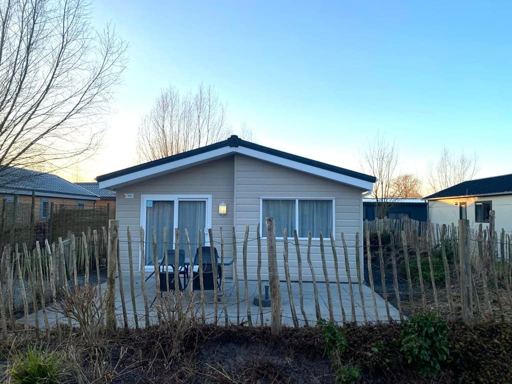 Houten vakantiewoning met een omheining van houten palen en een betegeld terras, omgeven door lage beplanting.