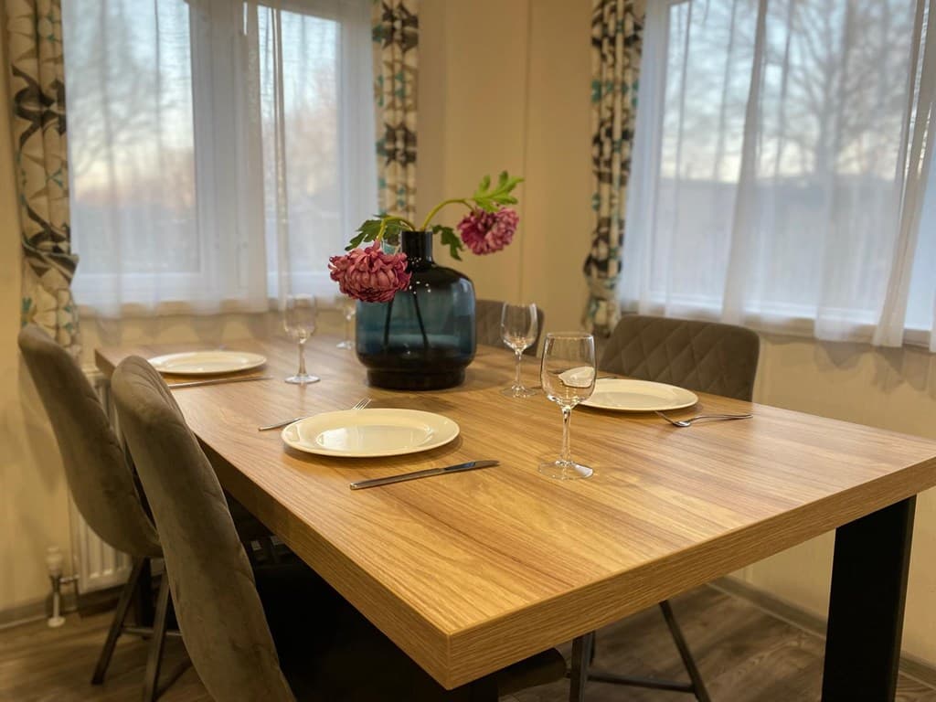 Eettafel met vier borden en glazen, een vaas met bloemen centraal, omgeven door lichtinval van de ramen.