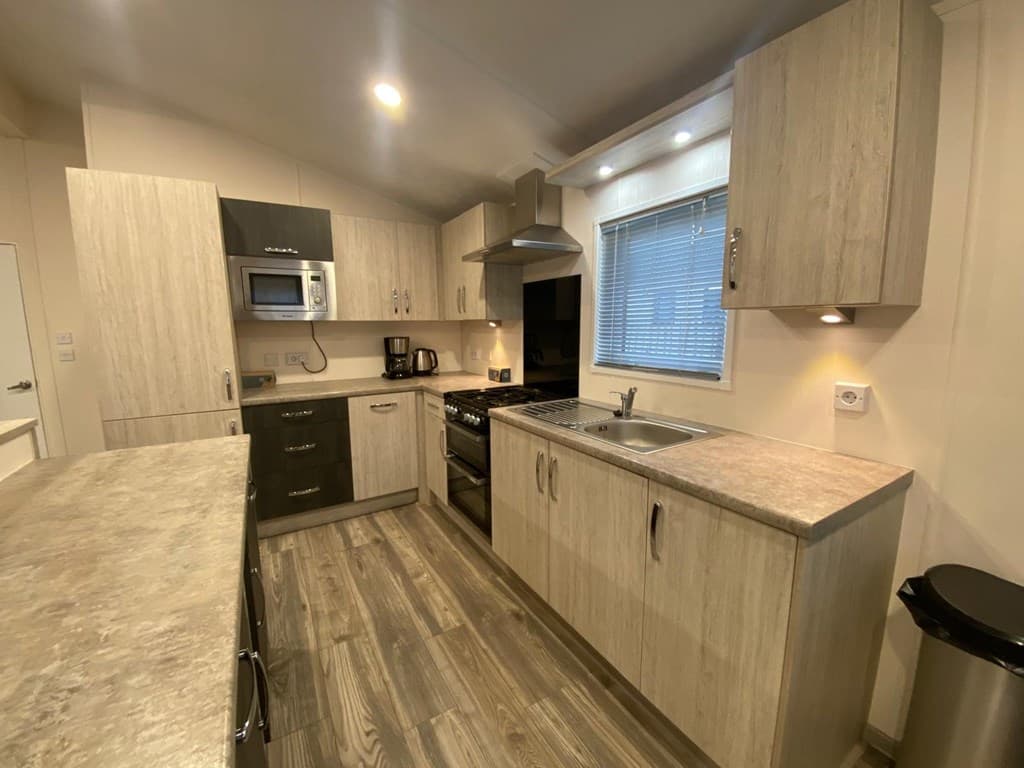 Moderne keuken met houten kasten, kookplaat, oven, magnetron en spoelbak met raam en verlichting boven het aanrecht.