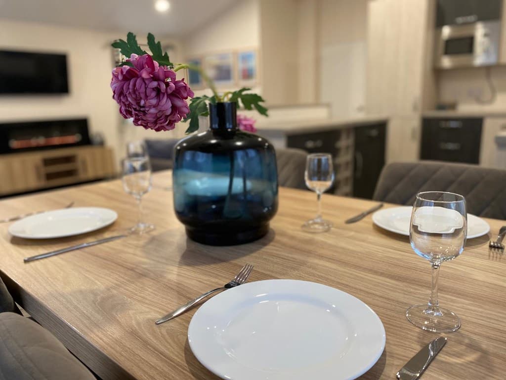 Eettafel met vier borden en glazen, een grote vaas met paarse bloemen en een moderne keuken op de achtergrond