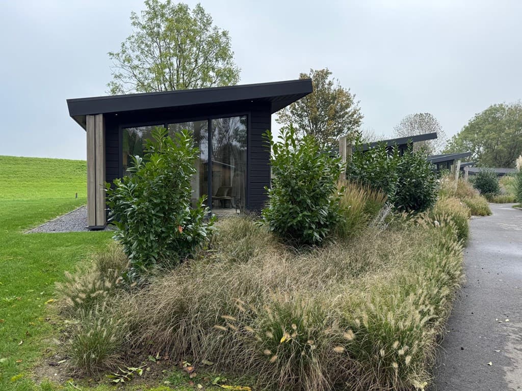 Moderne vakantiewoningen met grote ramen, omgeven door groen en struiken, gelegen aan een rustige laan.