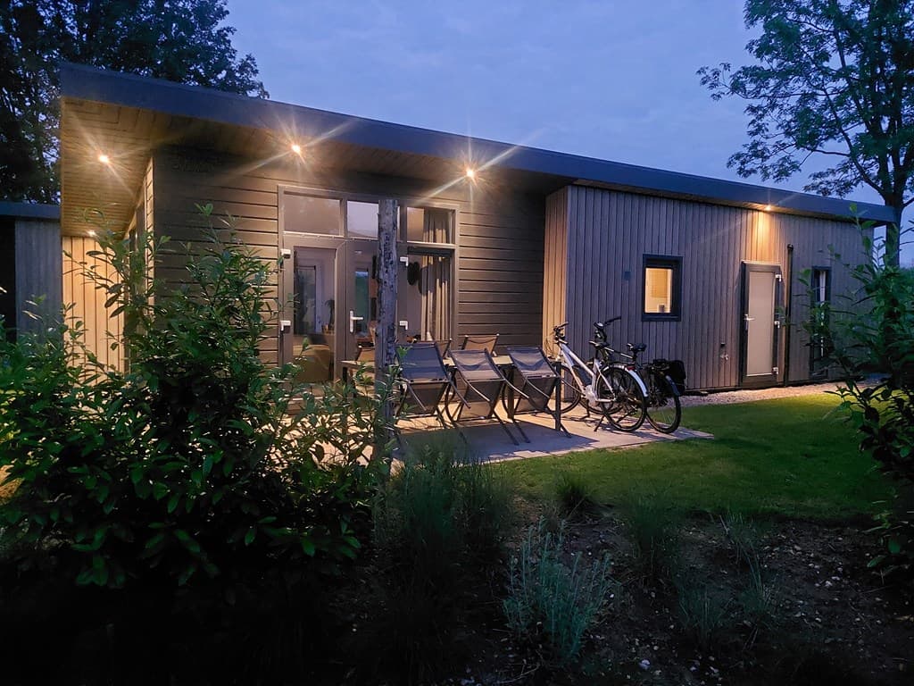 Moderne vakantiewoning met verlichting en terras, omgeven door bomen en een grasveld met fietsen.