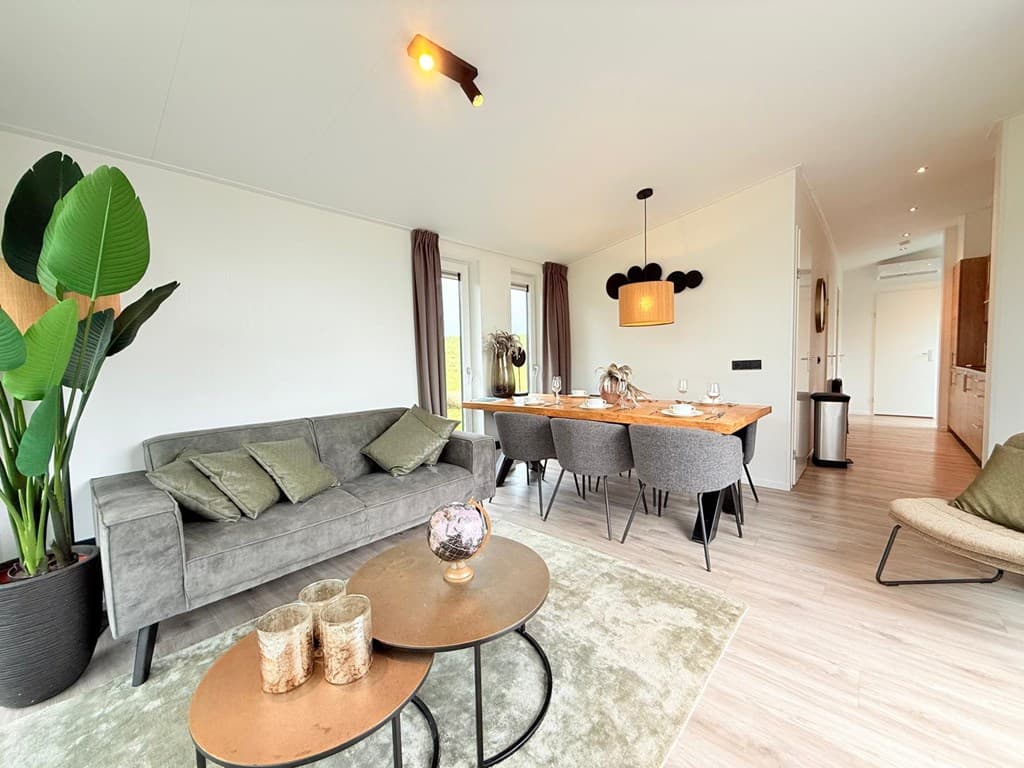 Woonkamer met een grijze bank, eettafel met stoelen en grote planten in een lichte, moderne inrichting