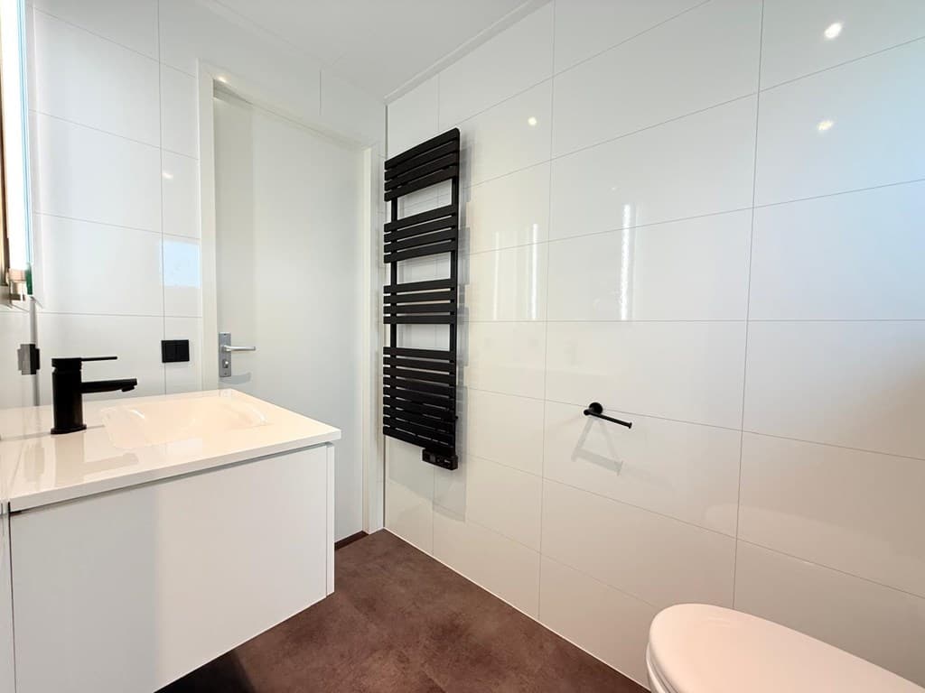 Moderne badkamer met witte tegels, wastafel, zwarte kranen en handdoekradiator tegen de muur.