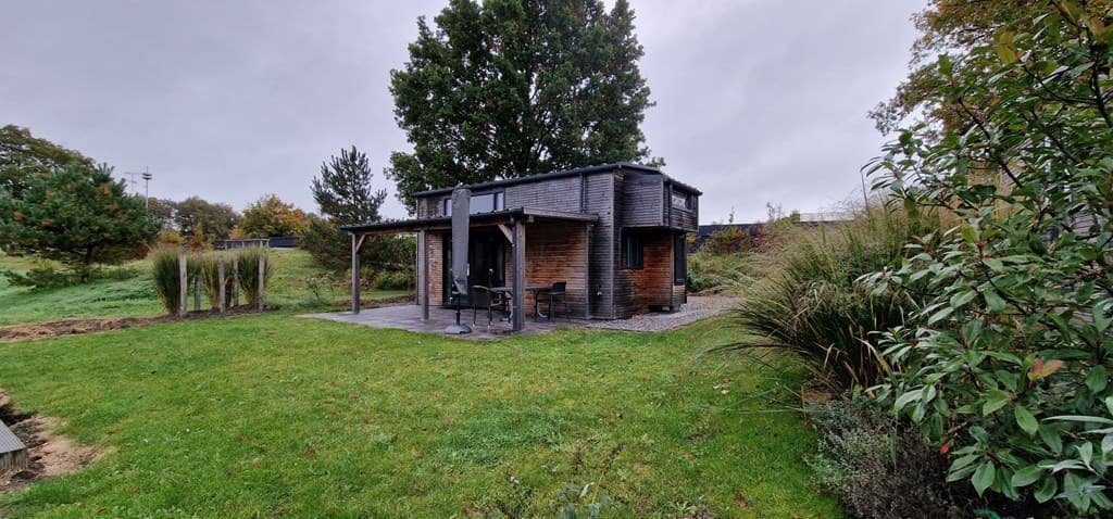 Zicht op een houten vakantiehuis met terras en tuin, omgeven door groen en enkele bomen.