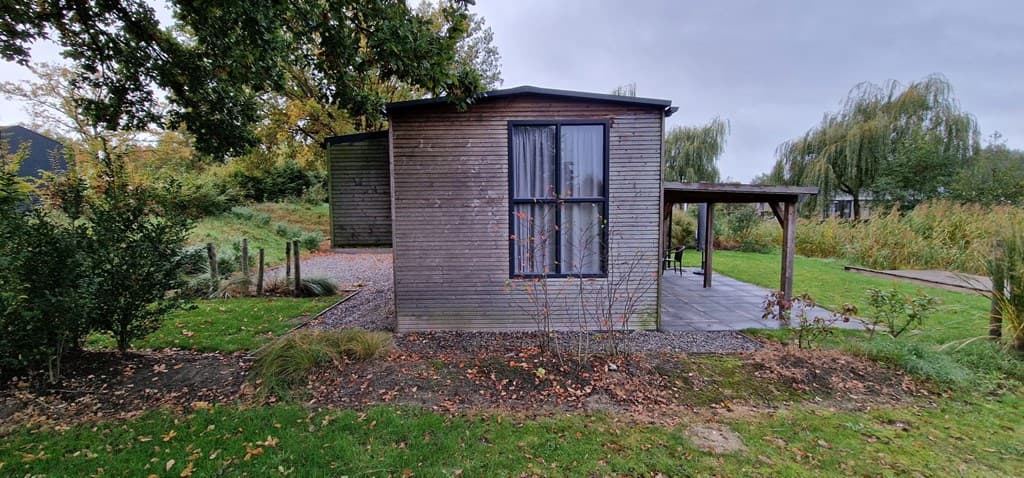 Houten vakantiewoning met grote ramen en overdekt terras in een groene omgeving met gras en bomen.