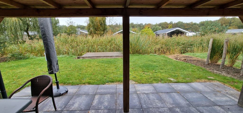 Zicht vanuit de overdekte terrasruimte op een groene tuin met gras en riet, omringd door bomen en woningen.