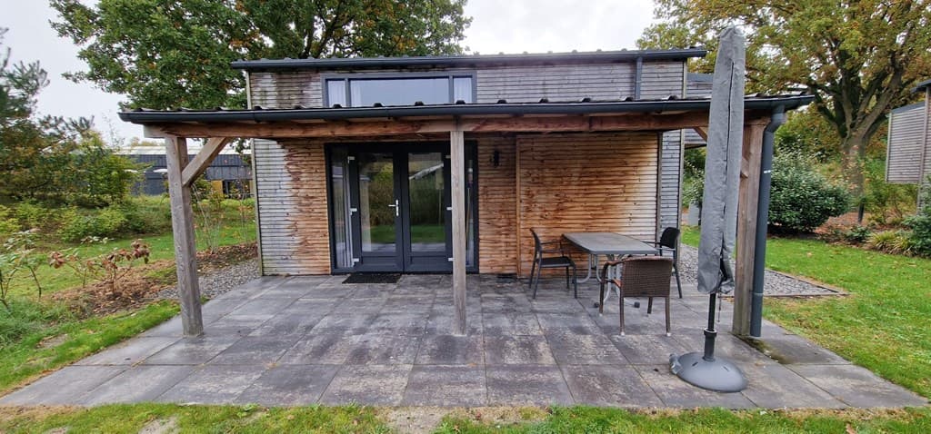 Voorzijde van een vakantiewoning met houten overkapping, terrasje en tuinmeubilair op een betegeld terras.