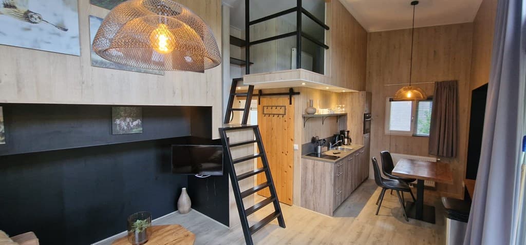 Ruime woonkamer met open keuken, trap naar een vide, houten meubels en een grote lamp boven de eettafel