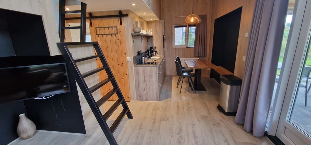 Moderne woonkamer met open keuken, houten accenten en een trap naar een vide, naast een eettafel en glazen deur naar buiten.