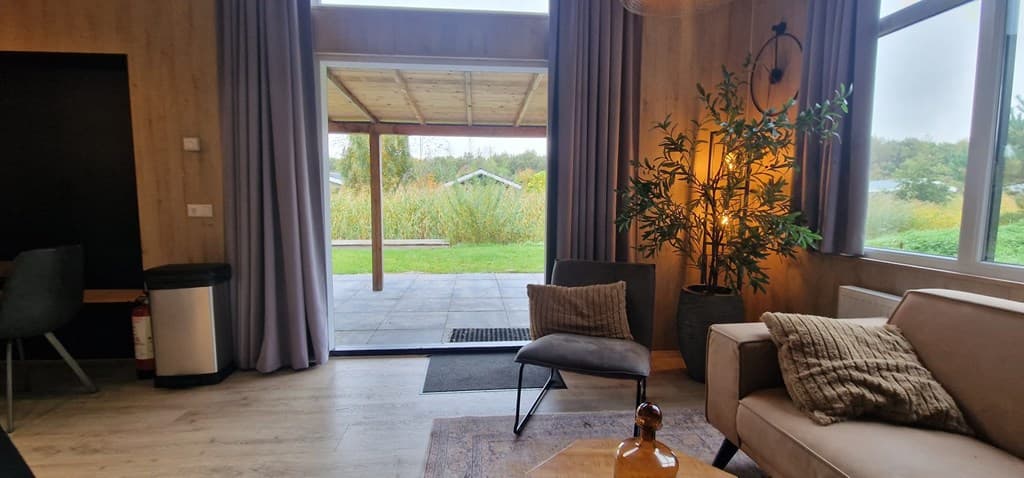 Knusse woonkamer met grote ramen, een bank, een plantenbak en toegang tot een overdekt terras met uitzicht op groen.