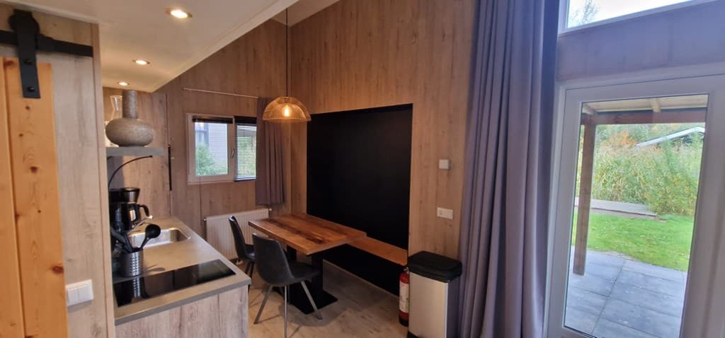 Moderne keuken met houten eettafel, stoelen en grote ramen met uitzicht op groene natuur buiten.
