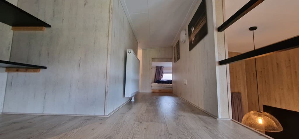 In een lichte gang met houten vloer en radiator, zicht op een slaapkamer aan het einde van de ruimte.