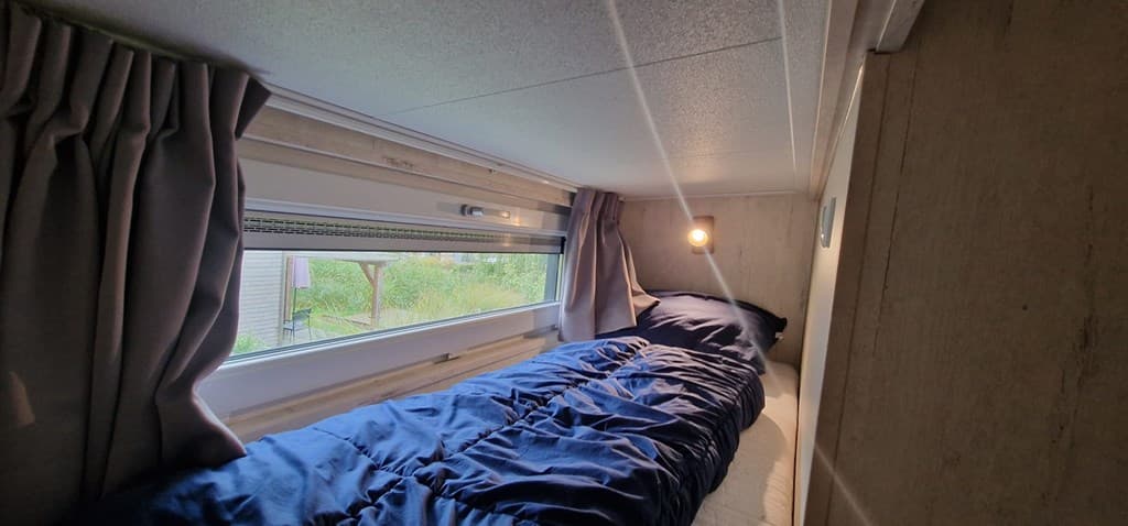 Slaapgedeelte met een éénpersoonsbed, ramen met gordijnen en zacht licht aan de wand.