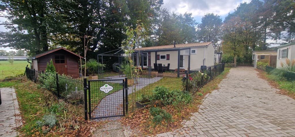 Tuin met terras en tuinmeubilair, hekwerk en houten schuur in een groene omgeving met bomen.