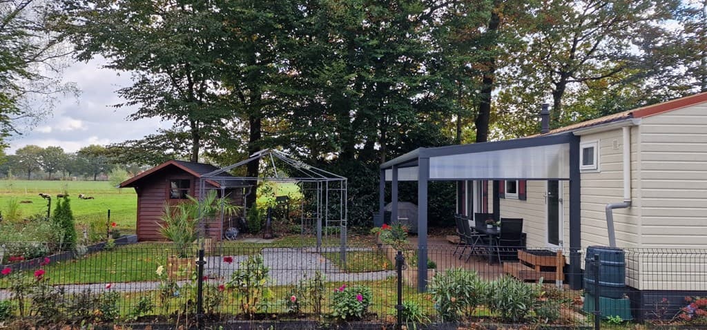 Tuin met gazon, pergola, houten schuur en bankje, omringd door bomen en een groene weide op de achtergrond